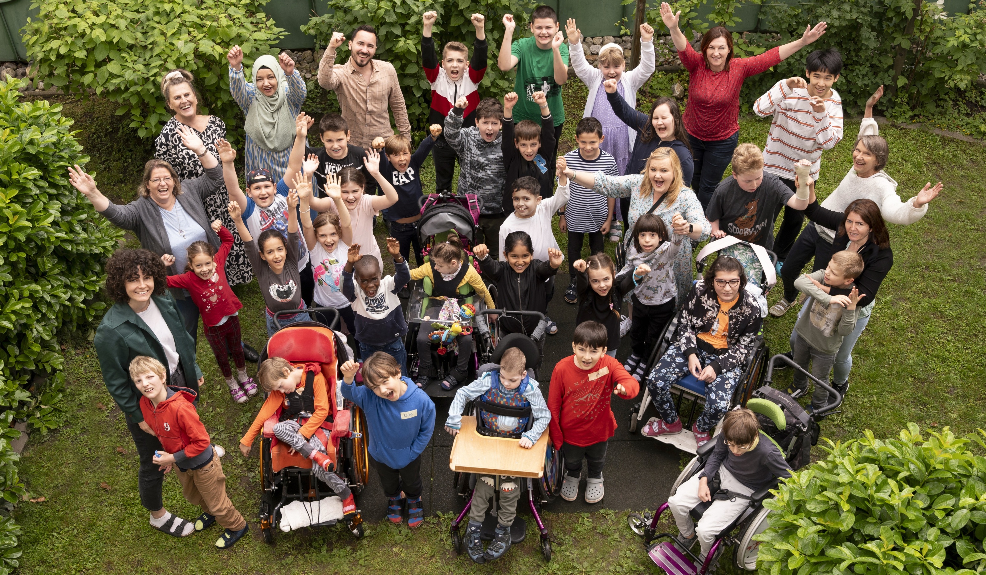 Kindern und Erwachsenen im Garten, einige sitzen in Rollstühlen. Alle heben die Hände in die Luft.